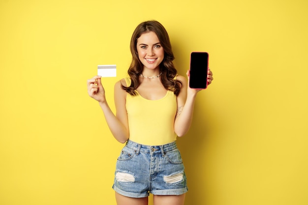 스마트폰 빈 전화 화면과 신용 카드를 보여주는 젊은 아름다운 여성 모델, 여름 준비, 탱크 탑과 반바지를 입고 노란색 배경 위에 서 있습니다.