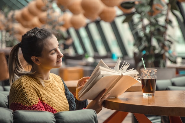 Молодая красивая женщина в оранжевом свитере читает интересную книгу в кафе