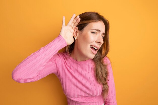 Young beautiful woman in pink top holding hand over her ear trying to listen to gossips 