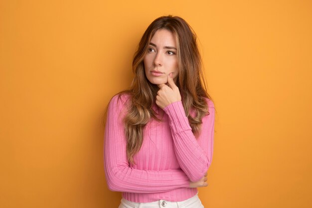 Young beautiful woman in pink top looking aside with pensive expression thinking standing over orange