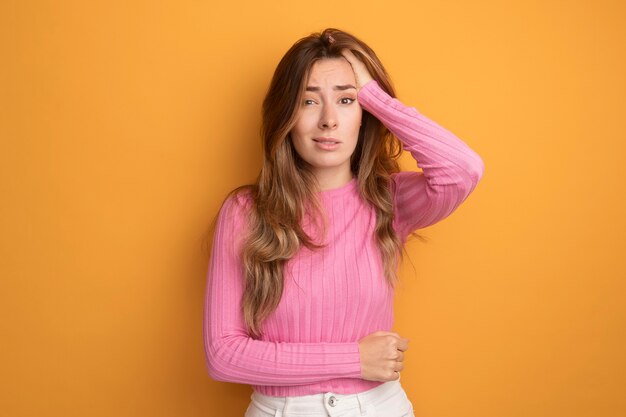 Young beautiful woman in pink top looking at camera confused and very anxious with hand on her head standing over orange background
