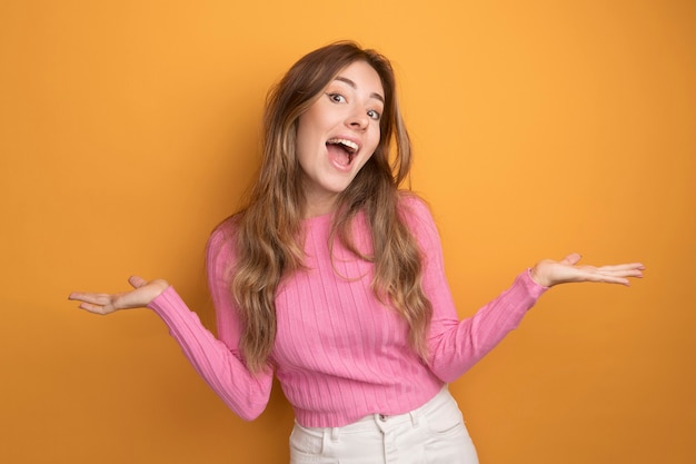 Young beautiful woman in pink top looking at camera happy and excited spreading arms to the sides