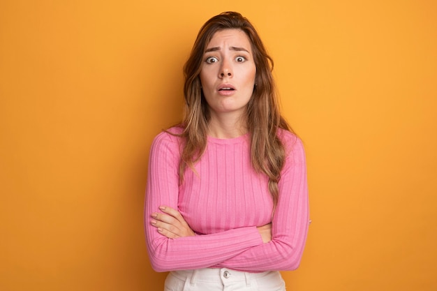 Young beautiful woman in pink top looking at camera surprised and confused standing over orange