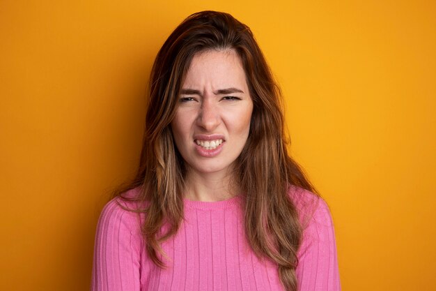 Young beautiful woman in pink top looking at camera with angry face standing over orange background