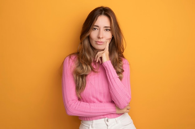 Young beautiful woman in pink top looking at camera with confident expression with hand on chin thinking standing over orange background