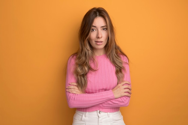 Young beautiful woman in pink top looking at camera with serious confident expression standing over orange background
