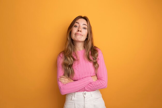Young beautiful woman in pink top looking at camera with skeptic expression on face standing over orange