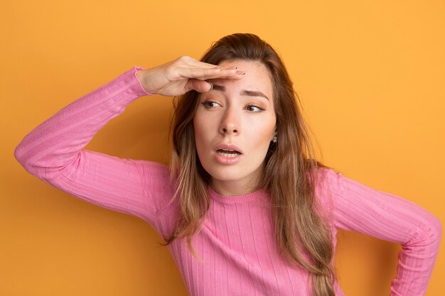 オレンジ色の上に立っている頭の上に手で遠くを見ているピンクのトップの若い美しい女性