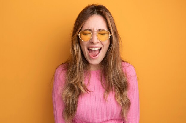 Young beautiful woman in pink top wearing glasses happy and excited screaming standing over orange