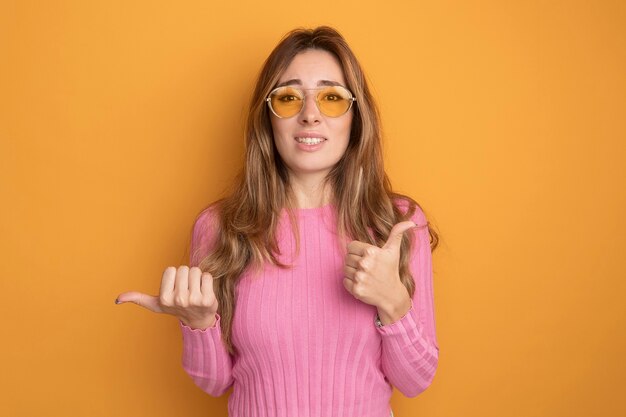 Young beautiful woman in pink top wearing glasses looking at camera confused showing thumbs up pointing to the side with thumb standing over orange background