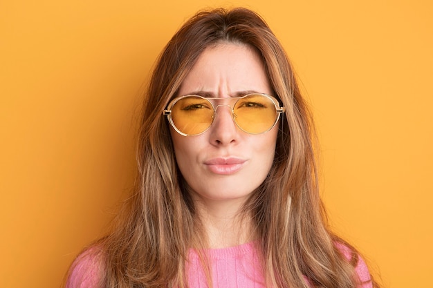 Young beautiful woman in pink top wearing glasses looking at camera frowning standing over orange