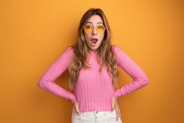 Young beautiful woman in pink top wearing glasses looking at camera surprised and amazed 