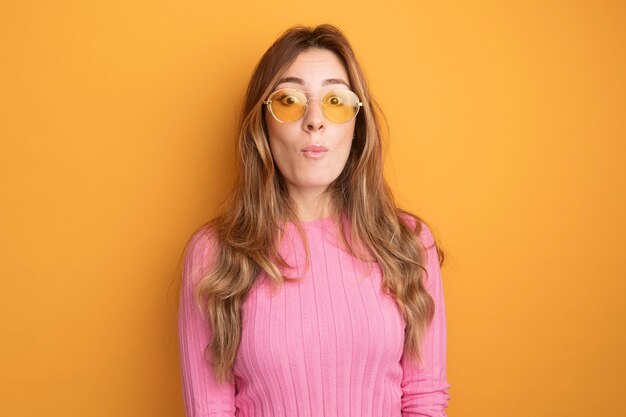 Young beautiful woman in pink top wearing glasses looking at camera surprised 