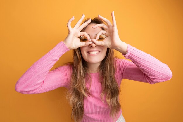Young beautiful woman in pink top wearing glasses looking through fingers making binocular gesture smiling standing over orange background