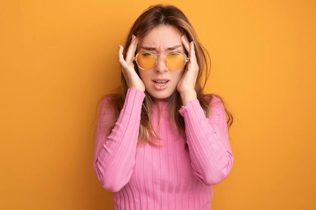 Young beautiful woman in pink top wearing glasses looking unwell touching her temples having headache standing over orange