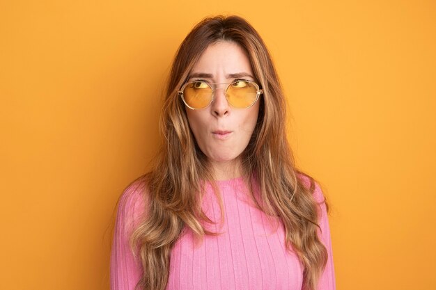 Young beautiful woman in pink top wearing glasses looking up puzzled standing over orange