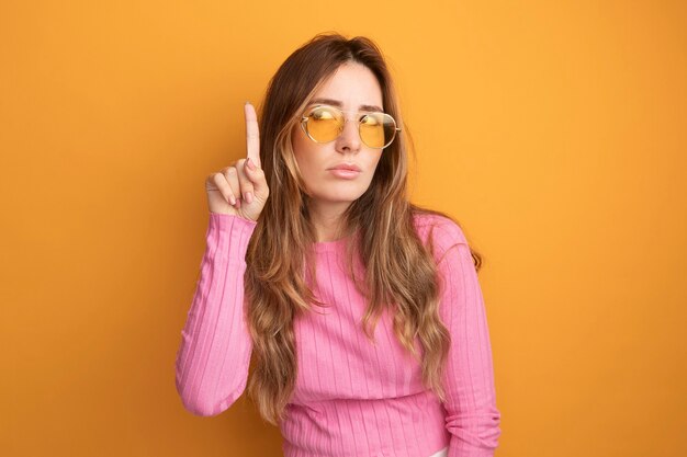 Young beautiful woman in pink top wearing glasses looking up with serious face showing index finger