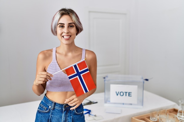 政治運動選挙でノルウェーの国旗を持った若い美しい女性が前向きで幸せそうに見え、歯を見せて自信に満ちた笑顔で微笑んでいる