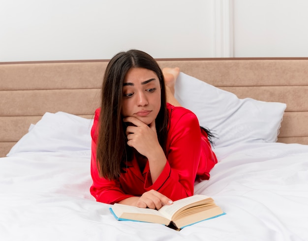 Young beautiful woman in red pajamas laying on bed with book reading looking confused in bedroom interior on light background