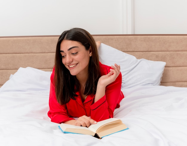 Young beautiful woman in red pajamas laying on bed with book relaxing enjoying weekend smiling with happy face in bedroom interior on light background