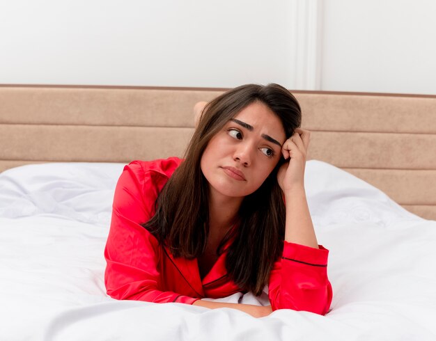 Молодая красивая женщина в красной пижаме, лежа на кровати с грустным выражением лица в домашнем интерьере на светлом фоне