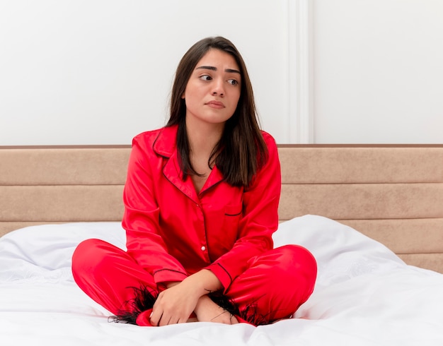 明るい背景の寝室のインテリアの顔に悲しい表情で脇を見てベッドに座っている赤いパジャマの若い美しい女性