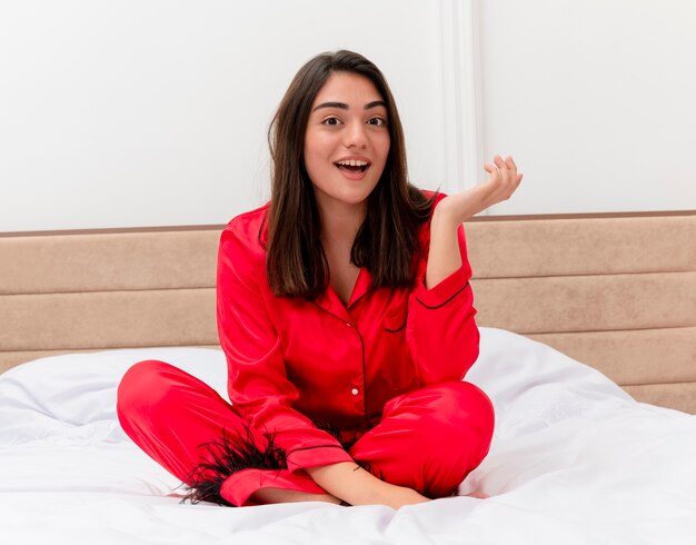 Young beautiful woman in red pajamas sitting on bed looking at camera happy and positive smiling cheerfully in bedroom interior on light background
