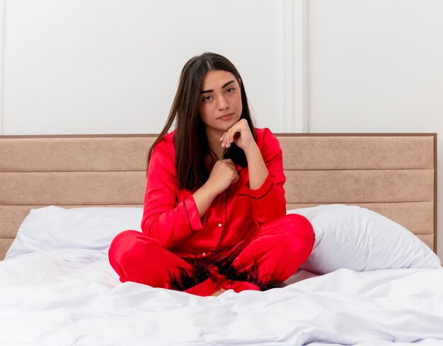 Молодая красивая женщина в красной пижаме сидит на кровати и смотрит в камеру с серьезным лицом в интерьере спальни на светлом фоне