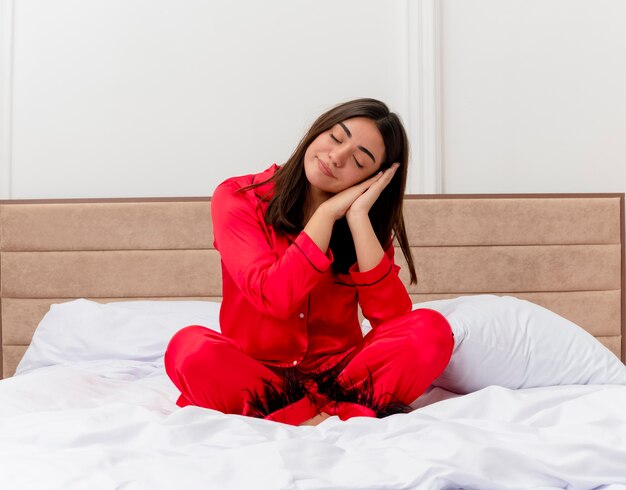 Young beautiful woman in red pajamas sitting on bed making sleep gesture holding palms together leaning head on palms in bedroom interior 