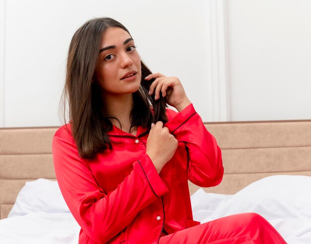 Молодая красивая женщина в красной пижаме сидит на кровати и разговаривает по мобильному телефону, уверенно глядя в интерьер спальни на светлом фоне