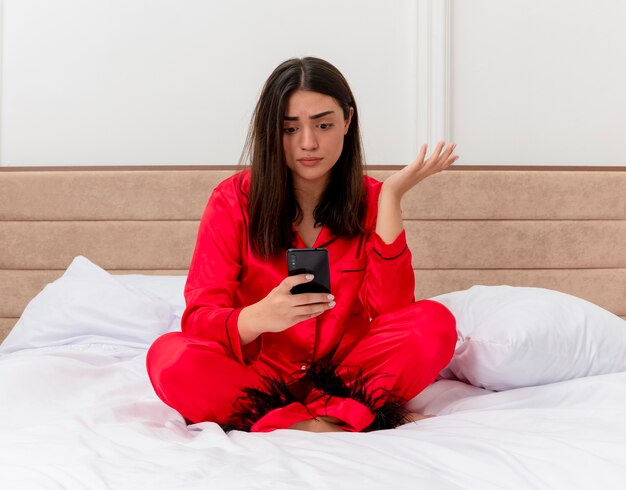 Молодая красивая женщина в красной пижаме, сидя на кровати с помощью смартфона, выглядит смущенной и недовольной в интерьере спальни на светлом фоне