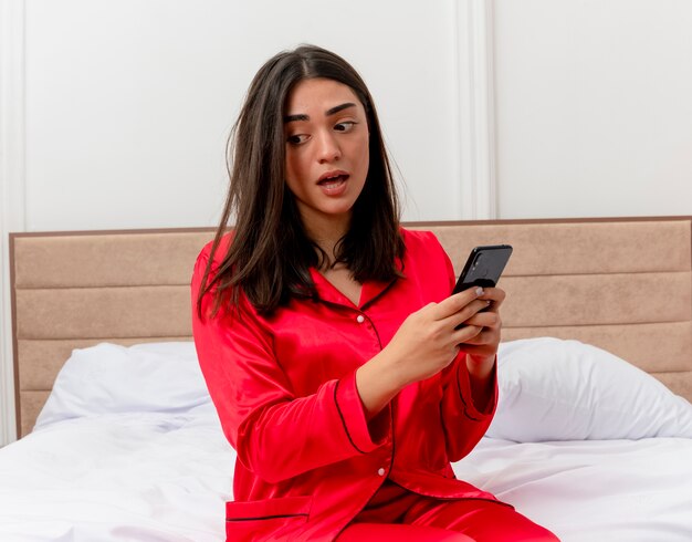 Young beautiful woman in red pajamas sitting on bed using smartphone looking confused and very anxious in bedroom interior on light background