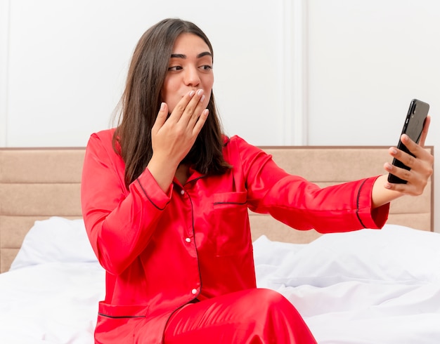 Young beautiful woman in red pajamas sitting on bed using smartphone looking at screen having video call blowing a kiss in bedroom interior on light background