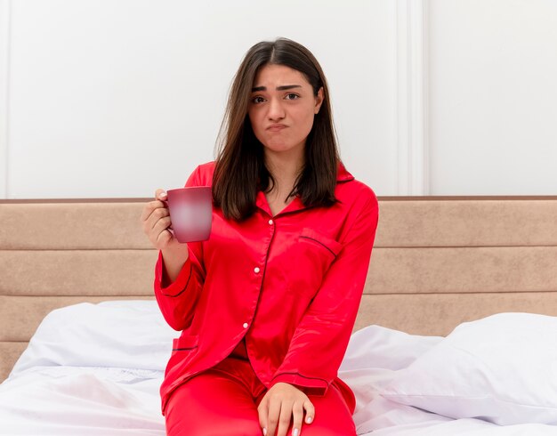 Young beautiful woman in red pajamas sitting on bed with cup of coffee looking at camera being confused and displeased in bedroom interior on light background