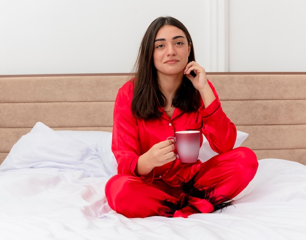 Young beautiful woman in red pajamas sitting on bed with cup of coffee looking at camera smiling in bedroom interior on light background