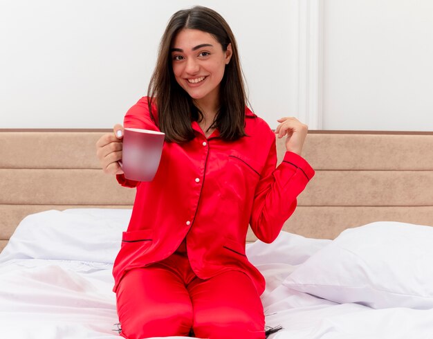 Young beautiful woman in red pajamas sitting on bed with cup of coffee looking at camera smiling cheerfully in bedroom interior on light background