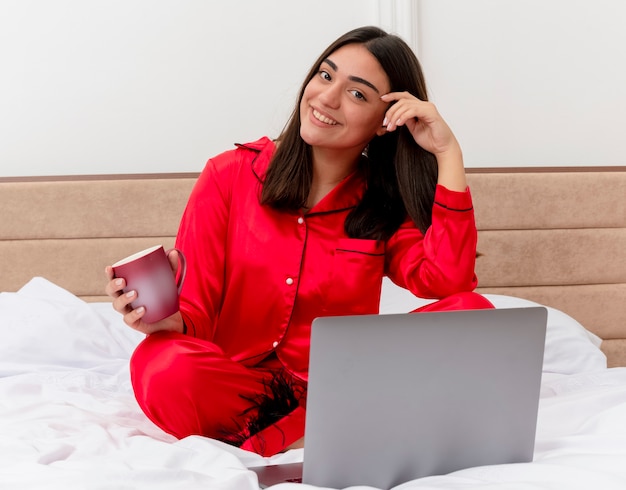 ラップトップとコーヒーのカップとベッドに座っている赤いパジャマの若い美しい女性は、明るい背景の寝室のインテリアで幸せでポジティブな顔に笑顔でカメラを見て