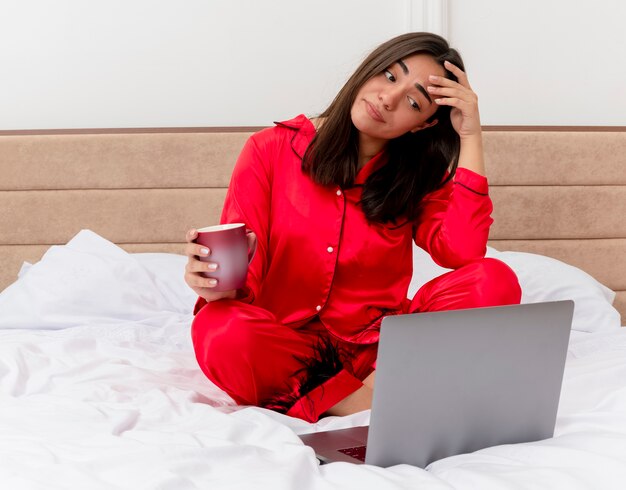 Молодая красивая женщина в красной пижаме, сидя на кровати с ноутбуком и чашкой кофе, выглядит смущенной и недовольной в интерьере спальни на светлом фоне
