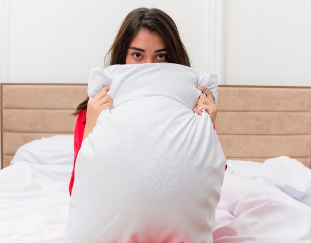 Young beautiful woman in red pajamas sitting in bed with pillow hiding behind it in bedroom interior 