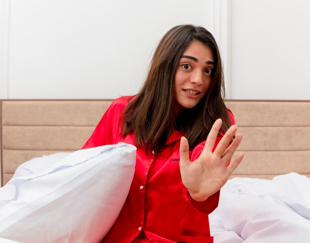 Young beautiful woman in red pajamas sitting in bed with pillow making stop gesture with hand being worried in bedroom interior on light background