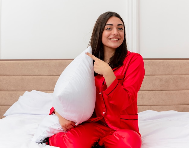 Молодая красивая женщина в красной пижаме сидит на кровати с подушкой, указывая указательными пальцами в сторону, весело улыбаясь в интерьере спальни на светлом фоне