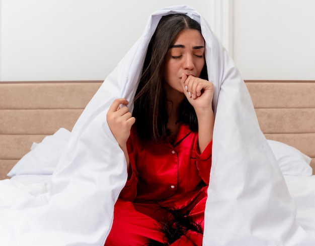 Young beautiful woman in red pajamas sitting on bed wrapping in blanket looking unwell coughing in bedroom interior on light background