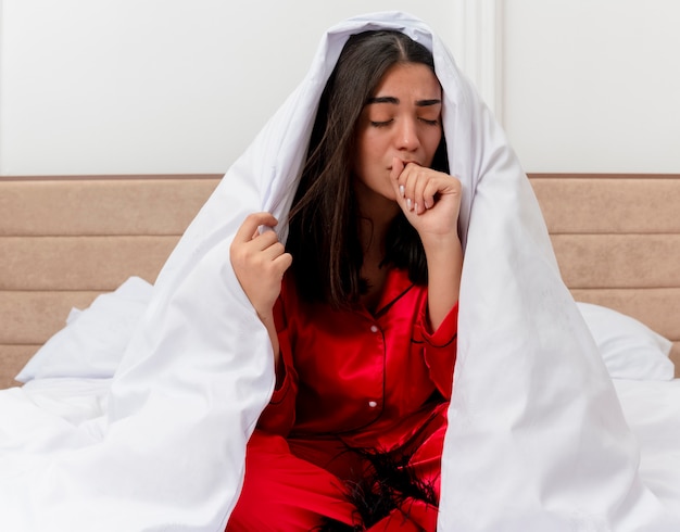 明るい背景の寝室のインテリアで体調不良の咳を探している毛布で包んでベッドに座っている赤いパジャマの若い美しい女性