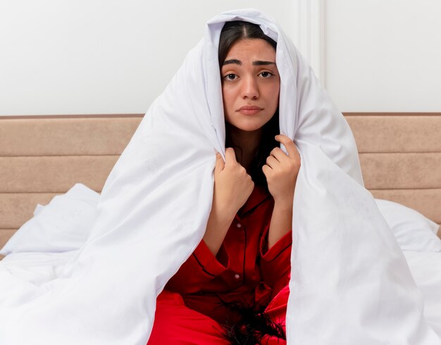Young beautiful woman in red pajamas sitting on bed wrapping in blanket looking unwell with unhappy face in bedroom interior on light background