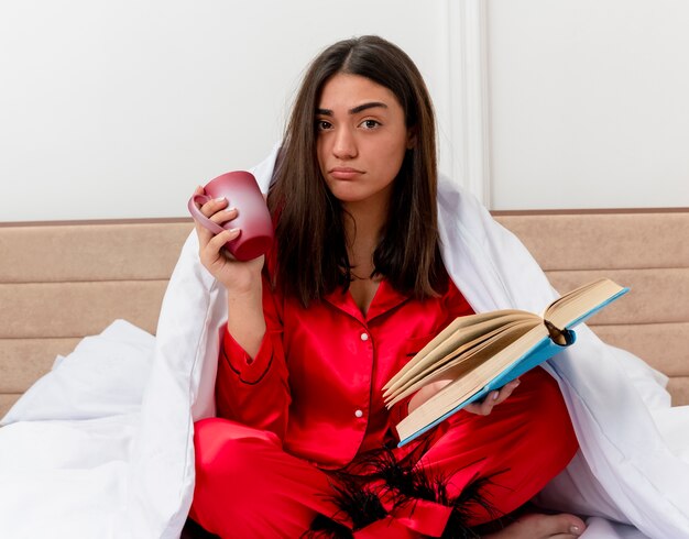 Young beautiful woman in red pajamas sitting on bed wrapping in blanket with cup of coffee and book looking at camera being displeased in bedroom interior on light background