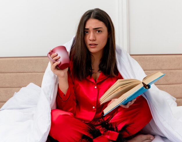 Young beautiful woman in red pajamas sitting on bed wrapping in blanket with cup of coffee reading book looking at camera being confused in bedroom interior on light background