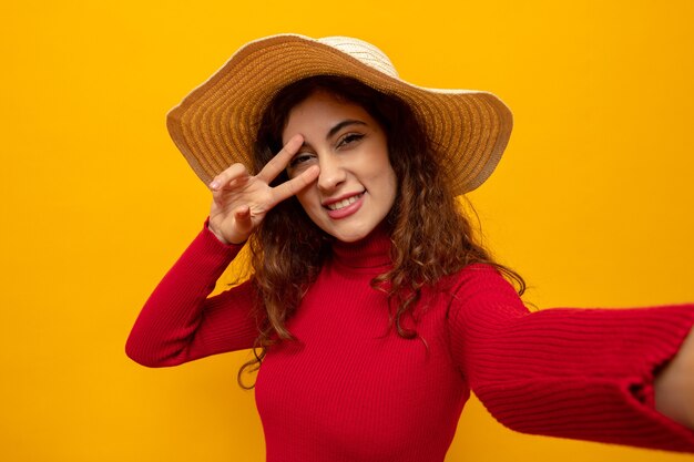 Young beautiful woman in red turtleneck in summer hat looking happy and cheerful smiling broadly showing v-sign