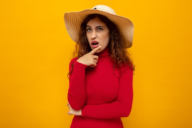 Young beautiful woman in red turtleneck in summer hat looking up with pensive expression standing on orange