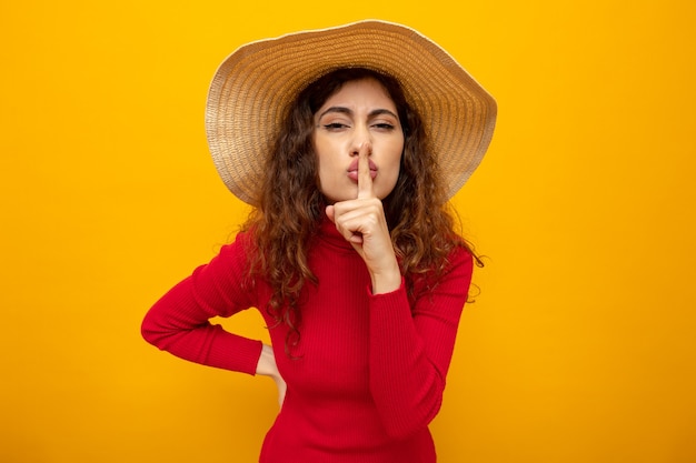 Young beautiful woman in red turtleneck in summer hat making silence gesture with finger on lips standing on orange