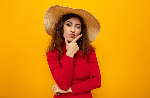 Young beautiful woman in red turtleneck in summer hat  with hand on her chin with pensive expression standing over orange wall